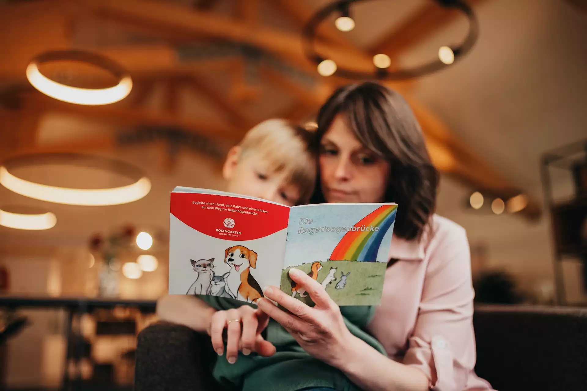 Frau und Kind lesen gemeinsam das Kinderbuch „Die Regenbogenbrücke“ mit Illustrationen von Hund, Katze und Hase.